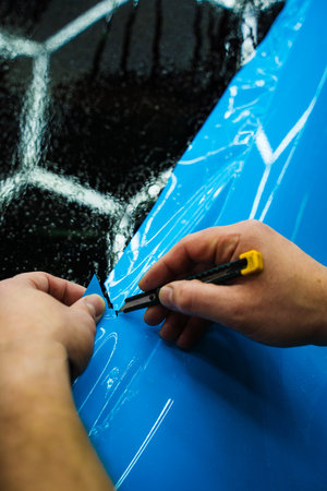 Cutting blue PPF with blade. Cutting blue paint protection film with blade during detailing job ensures precision edges. Detailing technician trims blue PPF on a car panel for perfect contour fitの写真素材