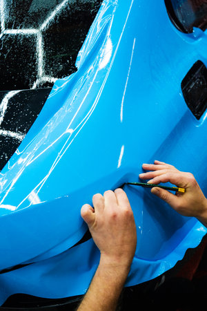 Cutting blue paint protection film with blade during detailing job ensures precision edges. Detailing technician trims blue PPF on a car panel for perfect contour fit. Cutting blue PPF with bladeの写真素材