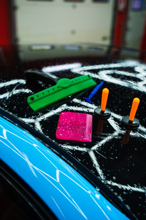 Car wrap tools, including squeegees and magnets on wet car hood. Car detailing tools to shape PPF wrap during installation for smooth coverage. PPF Paint Protection Film Tools on Car Hoodの写真素材