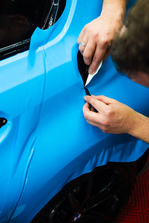 Detailing technician trims blue PPF on a car panel for perfect contour fit. Cutting blue PPF with blade. Cutting blue paint protection film with blade during detailing job ensures precision edgesの写真素材