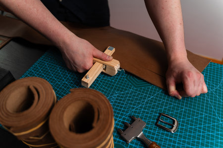 Handmade leather belt preparation with wooden strap cutter on cutting mat. Artisan is cutting...