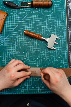Punching and detailing leather belt holes. Artisan punching holes into a leather strap for a handmade belt on a green cutting mat. Crafting belt holes with punch tool on leather strapの写真素材