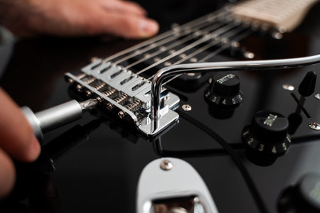 Technician fine-tunes the chrome bridge of a black electric guitar with a screwdriver. Chrome bridge...