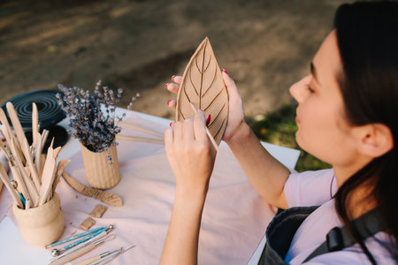 Young woman carves delicate leaf patterns into clay, showcasing her pottery skills. Artisan engraves a clay leaf with detailed lines. Woman Carving Clay Leafの写真素材