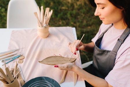 Artisan engraves a clay leaf with detailed lines. Woman Carving Clay Leaf. Young woman carves delicate leaf patterns into clay, showcasing her pottery skillsの写真素材