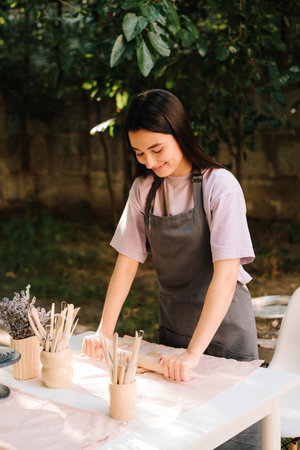 Young woman in an apron gently rolls out clay at outdoor pottery workshop. Girl prepares the clay...