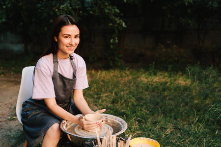 Young woman smiling while working with clay on pottery wheel. Girl shapes clay on the pottery wheel with smile. Smiling artisan shapes clay on the spinning pottery wheelの写真素材