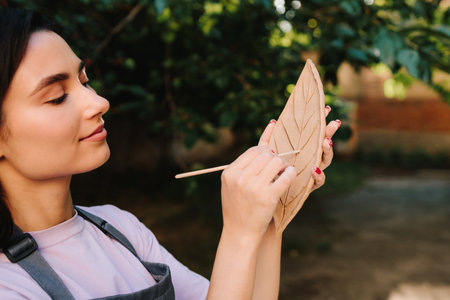Artisan engraves a clay leaf with detailed lines. Woman Carving Clay Leaf. Young woman carves delicate leaf patterns into clay, showcasing her pottery skillsの写真素材