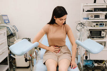 Abdominal discomfort during consultation signals possible gynecological issues. Women health concerns requiring gynecological care. Young woman sits in gynecological chair holding her abdomen in painの写真素材
