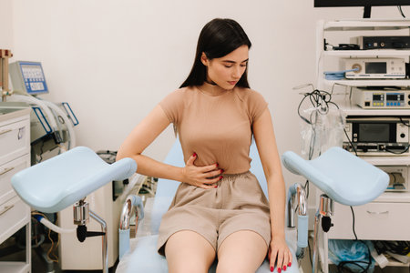 Young woman sits in gynecological chair holding her abdomen in pain. Abdominal discomfort during consultation signals possible gynecological issues. Women health concerns requiring gynecological careの写真素材