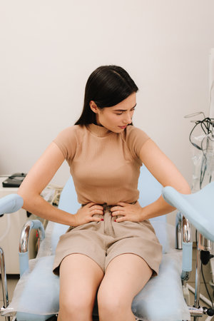 Young woman sits in gynecological chair holding her abdomen in pain. Abdominal discomfort during consultation signals possible gynecological issues. Women health concerns requiring gynecological careの写真素材