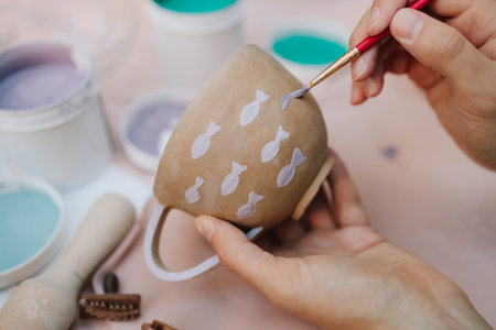 Ceramic painting of mug with fish pattern using brush strokes. Artist paints delicate purple fish on...