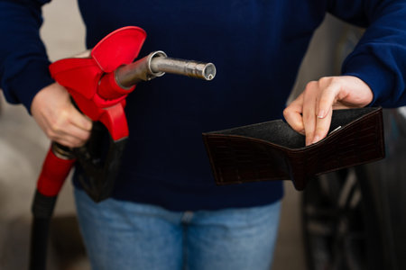 Hand holding empty wallet and gasoline nozzle at fuel station