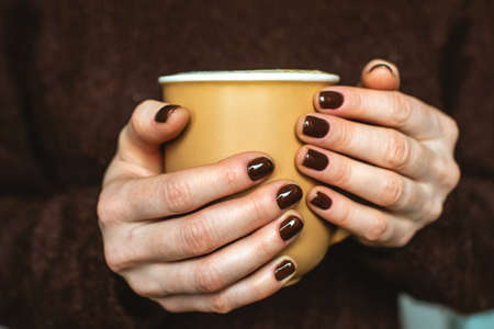 A woman wearing in a cozy warm sweater is holding a cup of aromatic coffee. The nails are coated with brown nail polish. Concept of elegance and stylish coffee manicure.の写真素材