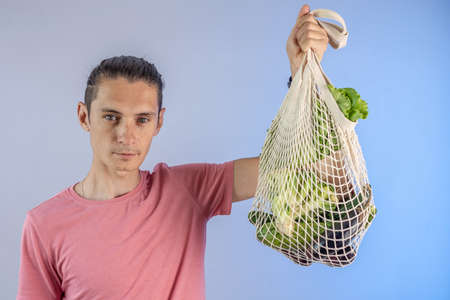 A modern young man is holding a string bag with purchases. Concept of eco-friendly behavior, the use of reusable bags and concern for the environment. Blue backgroundの写真素材