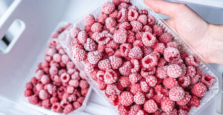 A person is taking a container of frozen raspberries out of the freezer of the fridge. Concept of frozen food, long term storage products.の写真素材