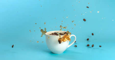 Coffee beans are falling into a white cup with black coffee and make splashes. Blue background.