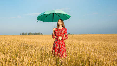 A young woman in a red dress and holding a green umbrella among the golden spikelets in a wheat field. Concept of protection, calmness and freedom.の写真素材