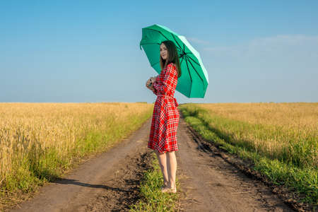 A young woman in a red dress and a green umbrella is walking along the road along a wheat field. Concept of protection, calmness and freedom.の写真素材