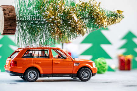 Toy orange car is carrying a Christmas tree on the roof against a background of paper pines. Concept of preparing for the holiday and deliveryの写真素材