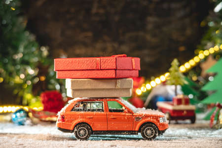 An orange toy car is going along a snow covered road against the background of festive lights and pines and carrying boxes with gifts. Concept of Christmas mood and preparation for the celebration.の写真素材