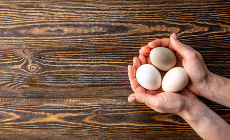Women's hands are carefully holding three raw organic eggs with shells of different unusual colors. Wooden background.の写真素材