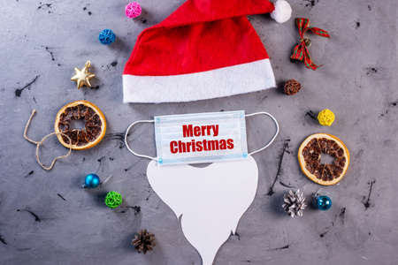 Red Santa hat, protective face mask with the inscription Merry Christmas and beard made of paper on a gray background with festive accessories.の写真素材