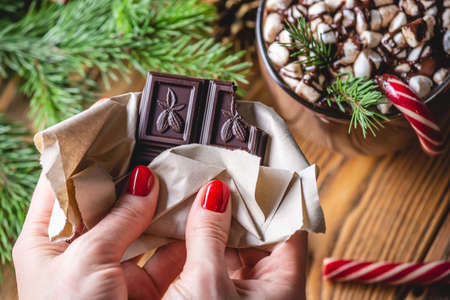 Women's hands are holding a chocolate bar on the background of a table on which there is a cup of...