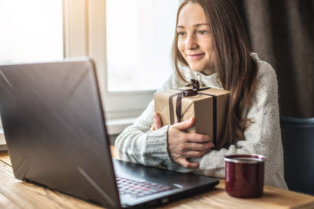 A happy girl in a sweater is sitting in front of a computer screen with a gift box in her hands. Concept of choosing gifts online and distance giving.の写真素材