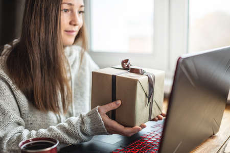 Woman in a sweater is sitting in front of a computer screen with a gift box in her hands. Concept of...