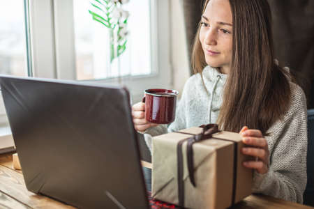 A young beautiful woman with a cup of coffee in her hands is sitting in front of a laptop screen and next to a gift box. Concept of online shopping gifts.の写真素材