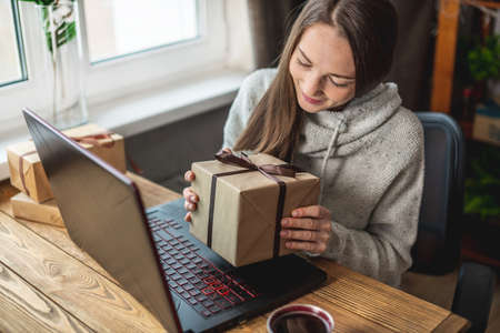 A happy girl in a sweater is sitting in front of a computer screen with a gift box in her hands. Concept of choosing gifts online and distance giving.の写真素材