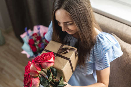 Happy young woman with a beautiful bouquet of fresh roses and a gift box in her hands at home. Concept of a holiday, romance, good and cozy mood in birthday.の写真素材