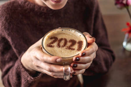 Woman in a knitted sweater is holding a glass cup of coffee in her hands. There are numbers 2021 on the cinnamon milk foam. Concept of a New year.の写真素材