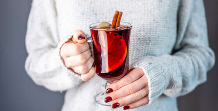 A woman in a white warm sweater is holding a transparent glass cup of mulled wine in her hand....