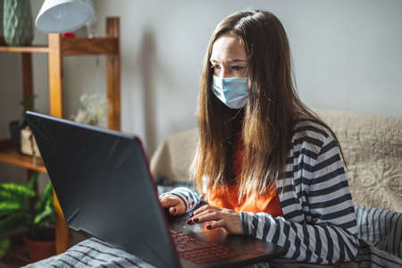 Young girl in a protective mask is sitting on a bed at home and using a laptop. Concept of distance...