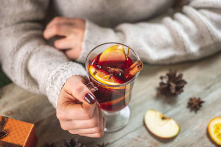 Woman's hands in a warm sweater are holding a cup of aromatic hot mulled wine on a wooden table....