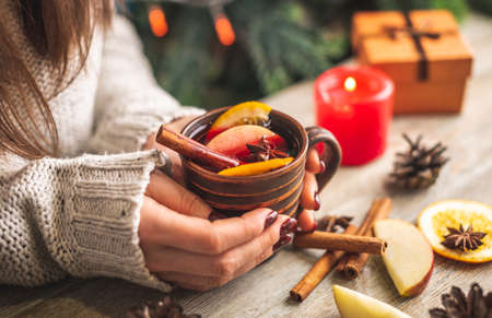 Woman's hands in a warm sweater are holding a cup of aromatic hot mulled wine on a wooden table....