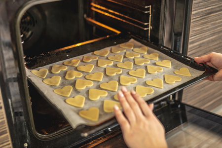 Woman is putting a baking sheet with delicious homemade cookies in the shape of a heart in the oven. Concept of fresh homemade cakes or a surprise for Valentine's Day.の写真素材