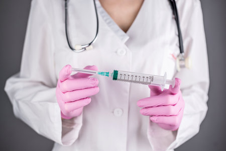 A female doctor in a medical gown and pink gloves is holding a syringe. Concept of health care and vaccination. Closeup and gray background.の写真素材