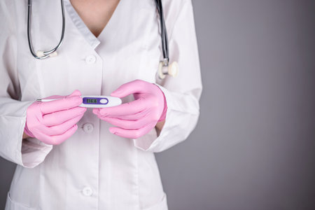 A female doctor in a white medical gown and pink gloves is holding a thermometer. Concept of health care. Closeup and gray background.の写真素材