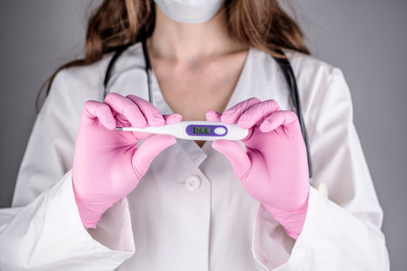 A female doctor in a white medical gown and pink gloves is holding a thermometer. Concept of health care. Closeup and gray background.の写真素材