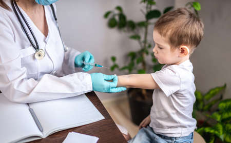 A young female doctor in a white medical coat, blue gloves, and a protective mask is vaccinating a small boy patient in his arm. Concept of health care and vaccination.の写真素材