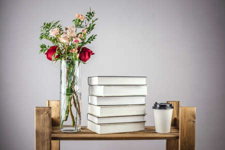 A vase of fresh beautiful flowers, a stack of books and a paper cup of coffee on a shelf of a wooden shelving. Concept of spring and a cozy pleasant atmosphere for reading and learning. Copy space.の写真素材