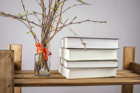 A vase with branches on which fresh green leaves bloom and a stack of books on a shelf of a wooden...