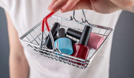 Woman is holding a miniature shopping basket with colorful bottles of nail polish. Closeup. Concept of choosing and buying nail polishes and accessories for manicure.の写真素材