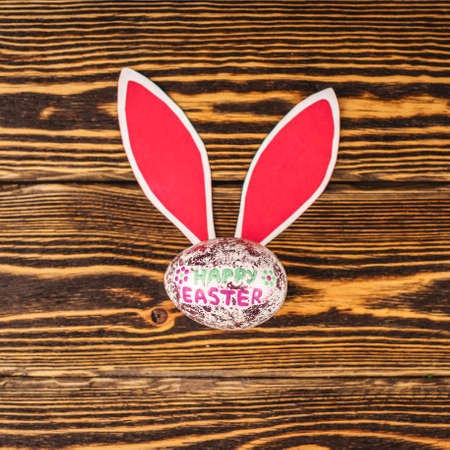 Shiny Easter egg with paper rabbit ears on wooden background. Top view.