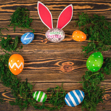 Bright colorful Easter eggs and paper rabbit ears on a wooden background with green moss. Top view...