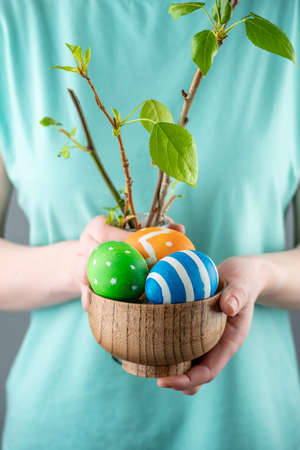 Woman is holding a wooden bowl with colorful Easter eggs and branches with the first green leaves of...