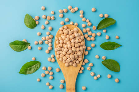 A portion of dried chickpeas in a wooden spoon on a blue table with green leaves. Concept of proper balanced nutrition and healthy vegetarian food.の写真素材
